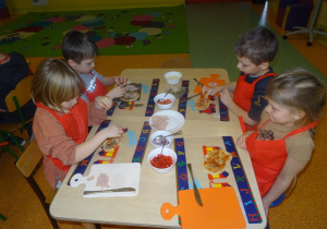Czwórka dzieci dekoruje szynką, warzywami i serem placek w kształcie koła.